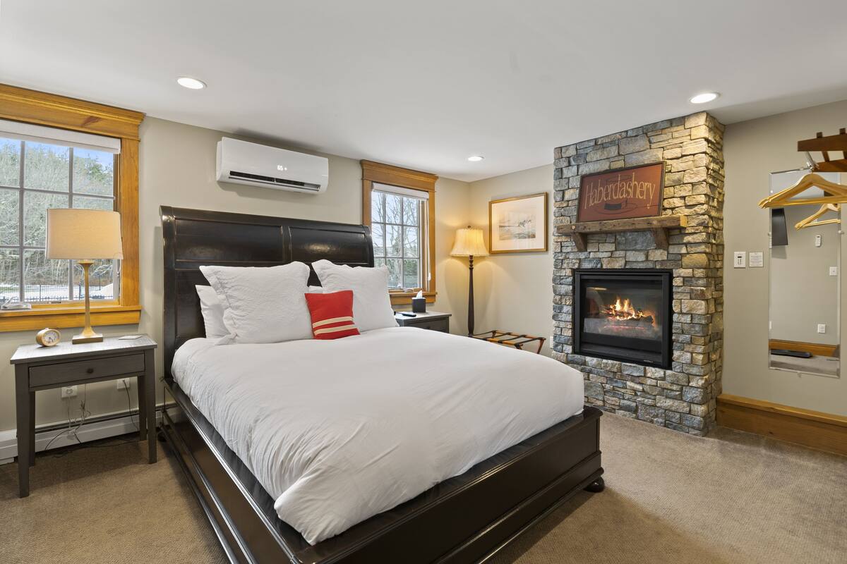 A bedroom with a large bed, a stone fireplace, and a window with a view of trees.