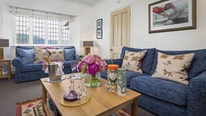A living room with two blue sofas, a wooden coffee table with decorative items, a vase of flowers, and a framed picture on the wall. - 4 Bed - Beach Cottage (Torquay)
