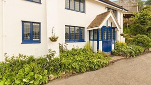A white building with blue trim and a blue door, surrounded by lush greenery. - 4 Bed - Beach Cottage (Torquay)