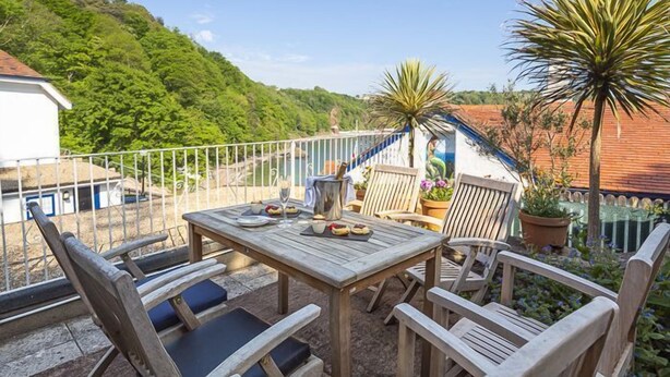 An outdoor patio with a wooden table and chairs, surrounded by potted plants and a view of a hillside. - 4 Bed - Beach Cottage (Torquay)