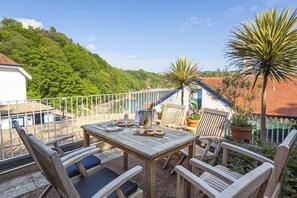 An outdoor patio with a wooden table and chairs, surrounded by potted plants and a view of a hillside. - 4 Bed - Beach Cottage (Torquay)