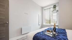 A modern bedroom with a bed, a bedside table with a bottle and glasses, a window with blinds, and a radiator. - Modern & Mellow 1-bed Apt w/ Free Parking & Wi-Fi (Bournemouth)