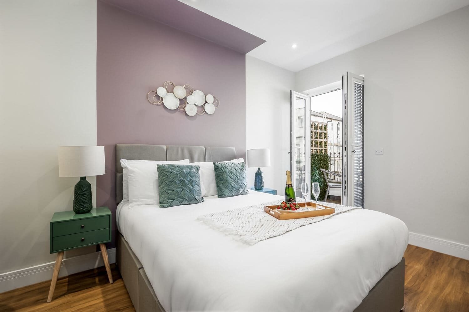 Quarto moderno com cama, mesas de cabeceira, um detalhe decorativo na parede e vista para o exterior.