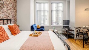 A modern hotel room with a brick accent wall, a large bed with orange pillows, a breakfast tray, a TV, and a dining area with a table and chairs. - Cosy, Beautiful Studio Apt w/ Free Parking & Wi-Fi (Bournemouth)