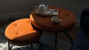 A room with a view of the sea, a small round table with a teapot and cup, and an orange ottoman.
