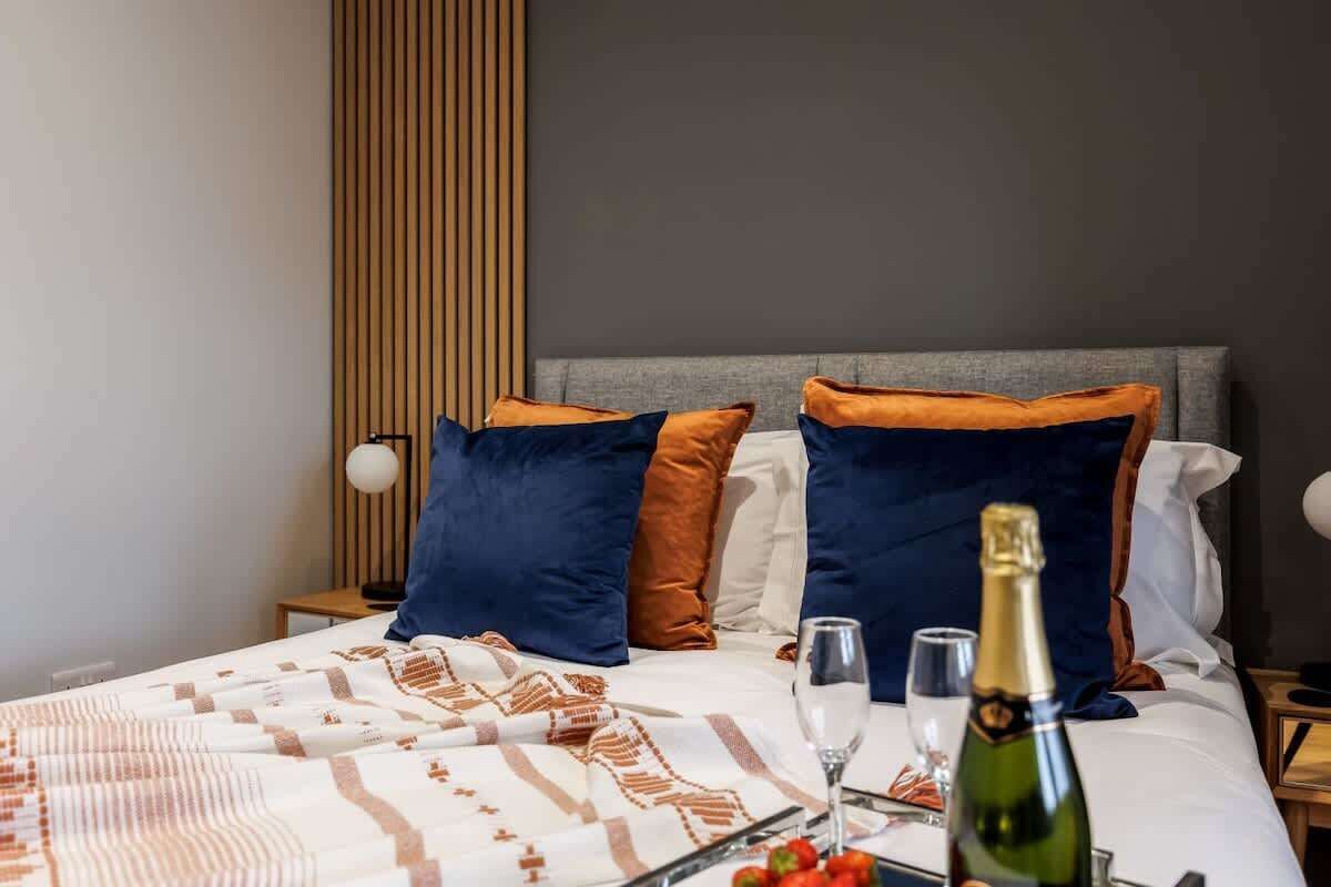 A neatly made bed with a patterned blanket, two pillows, a bottle of champagne, and glasses on a bedside table.