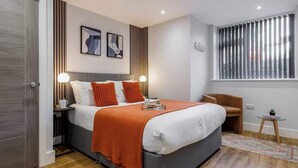 A modern bedroom with a large bed, bedside lamps, a small table, and a window with blinds. - Simply Beautiful 1-bed Apt w/ Free Parking & WiFi (Bournemouth)
