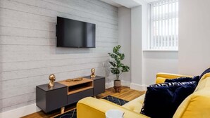 A modern living room with a flat-screen TV mounted on a wooden wall, a yellow sofa, a small side table with decorative items, and a potted plant. - Simply Beautiful 1-bed Apt w/ Free Parking & WiFi (Bournemouth)