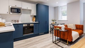 A modern kitchen with white and blue cabinetry, a built-in oven, and a microwave. There is a dining area with a table and orange sofa. - Comfy & Cosy 1-bed Apt w/ Free Parking & WiFi (Bournemouth)