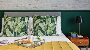 Un lit bien fait, avec deux oreillers ornés d’un motif de feuilles tropicales, une table de chevet avec une lampe et une petite plante, et un plateau avec de la nourriture et des boissons posé sur le lit.