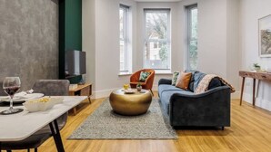 A modern living room with a sofa, coffee table, and TV. There are windows with blinds, a small table with a glass of wine, and a chair. - Spacious Seaside 2-Bed Apt w/ Free Gym & Parking (Bournemouth)
