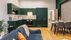 A modern kitchen with dark green cabinets, a dining area with a table and chairs, and a blue sofa with a throw blanket. - Spacious Seaside 2-Bed Apt w/ Free Gym & Parking (Bournemouth)