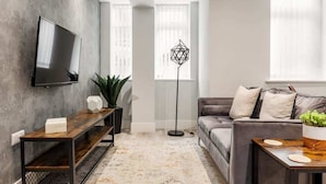 A modern living room with a flat-screen TV mounted on a concrete wall, a dark wooden bench, a grey sofa with cushions, and a floor lamp. - Spacious, Beautiful 1-Bed Apt; Central, Wi-Fi, PKG (Bournemouth)