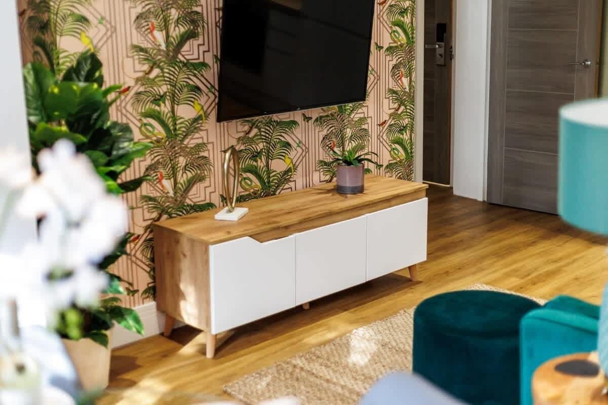 A modern living room with a wooden console table, a flat-screen TV mounted on a patterned wallpaper wall, and a teal accent chair.