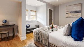 Un dormitorio moderno con cama, escritorio y silla. Se ve una ventana con persianas, un cuadro en la pared y una botella de champán sobre la cama.