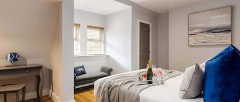 A modern bedroom with a bed, a desk, and a chair. There is a window with blinds, a painting on the wall, and a bottle of champagne on the bed.