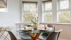 Un comedor con una mesa de vidrio, cuatro sillas grises y un jarrón con flores blancas.