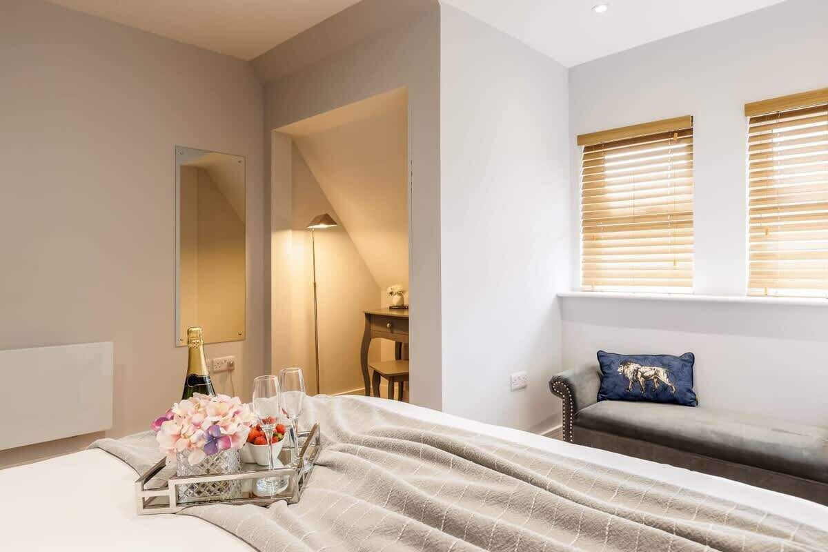 Ett hotellrum med en säng, ett sängbord med ett bricka innehållande champagne, glas och blommor; en soffa med en kudde; en stor spegel; och ett fönster med persienner.