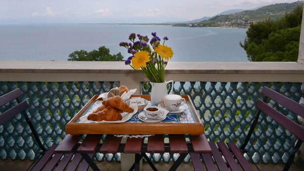 Varanda com uma bandeja de croissants, uma chávena de café e um vaso de flores, com vista para um corpo de água e uma cidade ao longe.