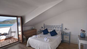 Ein Schlafzimmer mit einem großen Bett, zwei Nachttischen, einem kleinen Tisch und einem Stuhl. Durch eine Schiebetür bietet sich Meerblick.