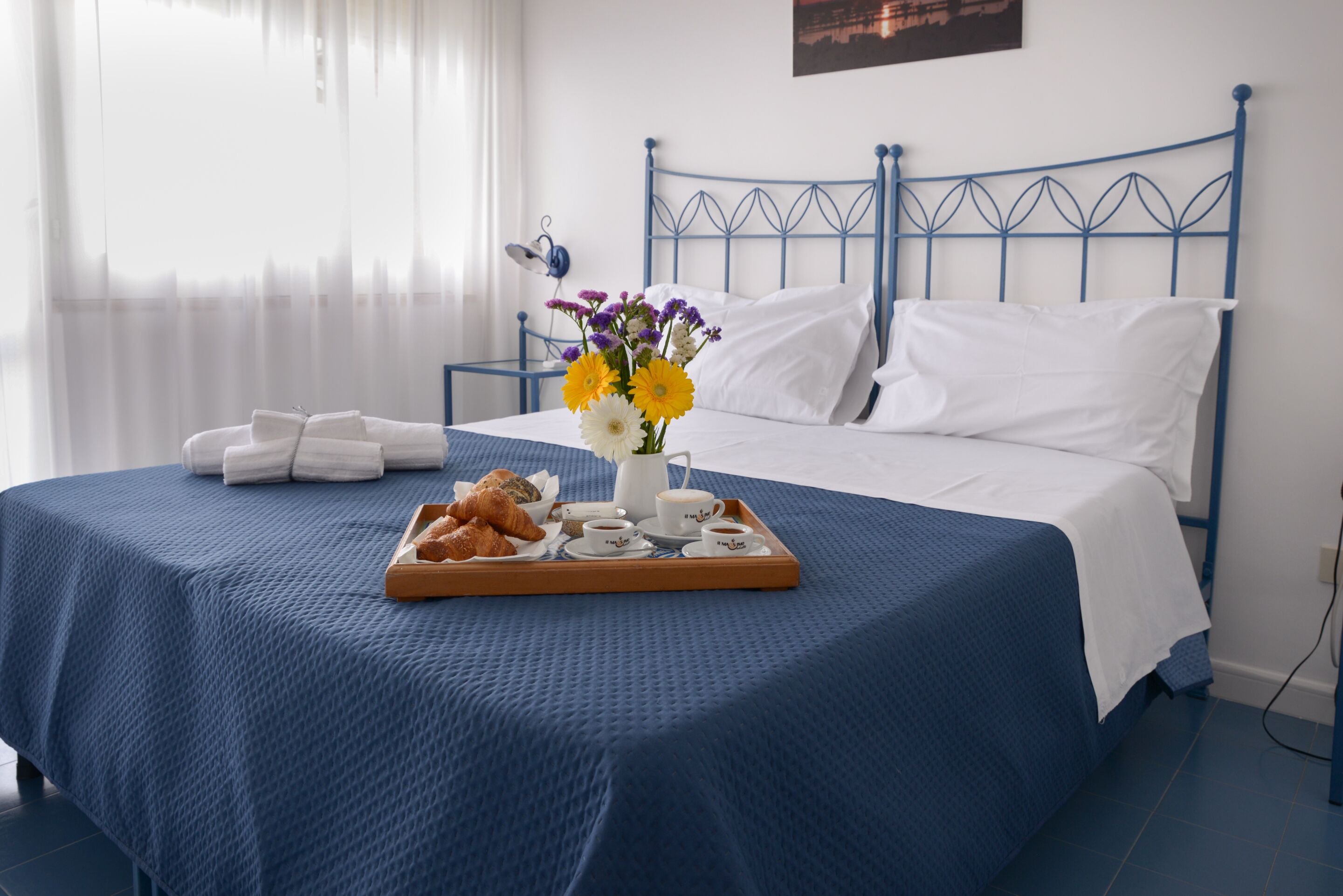 Un lit bien fait, avec une tête de lit bleue, un plateau de croissants, un vase de fleurs et une table de chevet avec une lampe.
