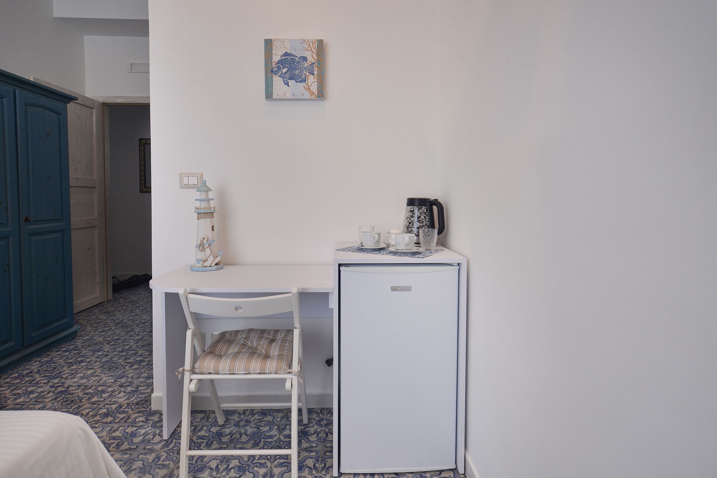 Una stanza piccola con una scrivania bianca, una sedia, un piccolo frigorifero bianco e un armadietto blu.