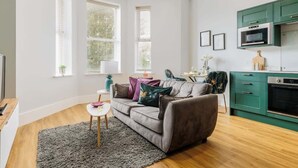 A modern living room with a grey sofa, a small white table, and a green kitchen area with a microwave and oven.