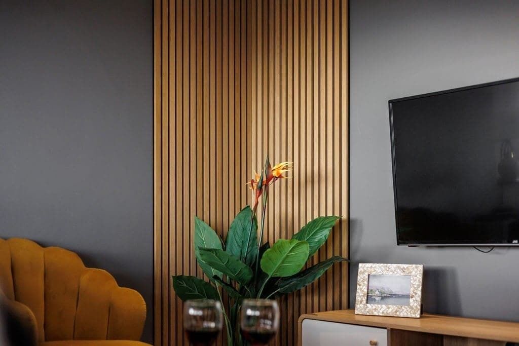 A room with a modern design, featuring a wooden slat wall, a flat-screen TV, a yellow armchair, a small table with a picture frame, and a potted plant.