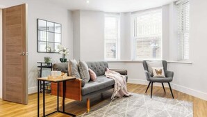 A modern living room with a grey sofa, a small table, and a chair. There are two windows with blinds, a mirror on the wall, and a door. - Spacious Garden, 2-Bed Apt w/ Free Gym & Parking (Bournemouth)