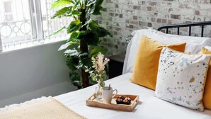 A neatly made bed with a white and yellow pillow, a tray with a cup and muffins, and a plant beside a window with a brick wall.