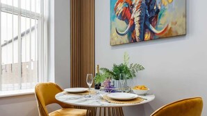 A dining area with a round table, two yellow chairs, a vase with plants, and a colorful abstract painting on the wall.