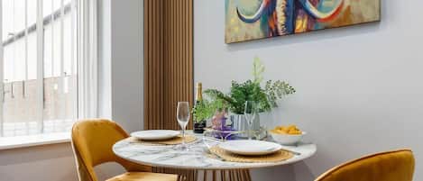 A dining area with a round table, two yellow chairs, a vase with plants, and a colorful abstract painting on the wall.
