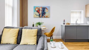 A modern living room with a grey sofa, yellow cushions, a coffee table, and a kitchen area with a grey cabinet and a window.