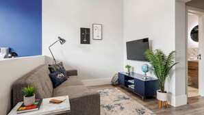 A modern living room with a grey sofa, a coffee table with books and a plant, a TV on a blue stand, and a large potted plant. - Comfy Seaside Studio Apt w/ Free Gym & Wi-Fi (Bournemouth)