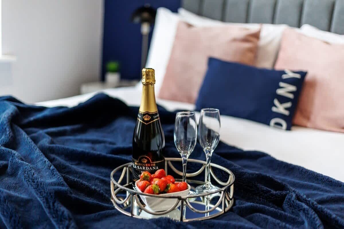 En säng med ett mörkblått täcke, en flaska champagne, två glas och ett fat med jordgubbar.