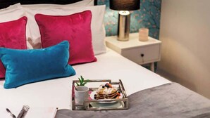 Un lit bien fait, avec deux oreillers, une table de chevet sur laquelle se trouvent une lampe et une bougie, et un plateau avec des crêpes et une tasse.