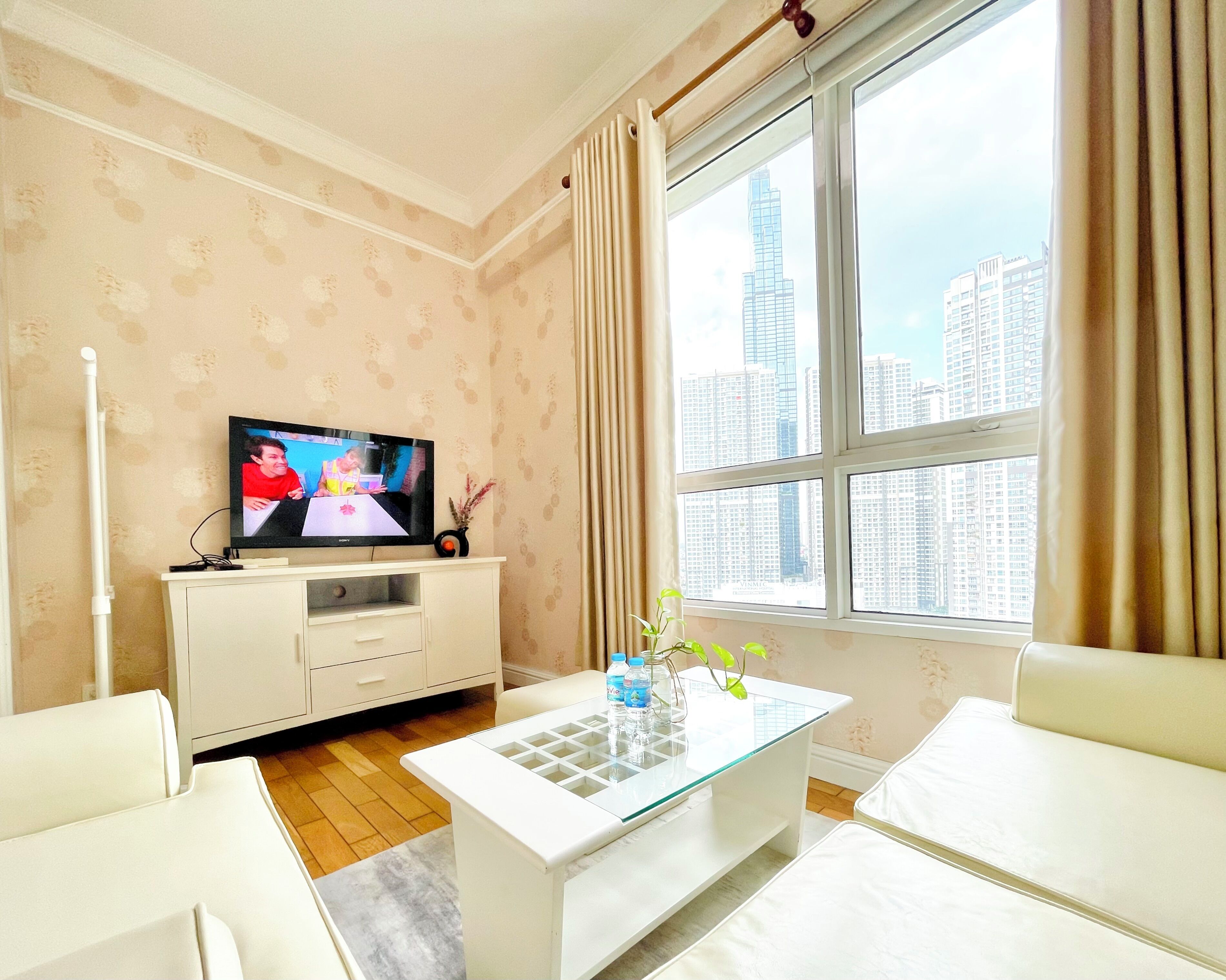 A modern living room with a television, white furniture, a glass coffee table, and a view of a cityscape through large windows.