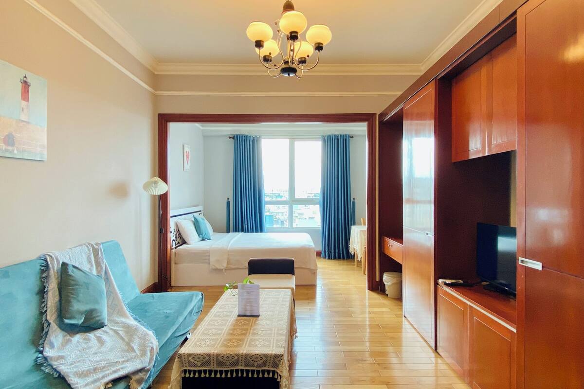 A hotel room with a bed, a sofa, a television, a chandelier, and a view of the city through the window.