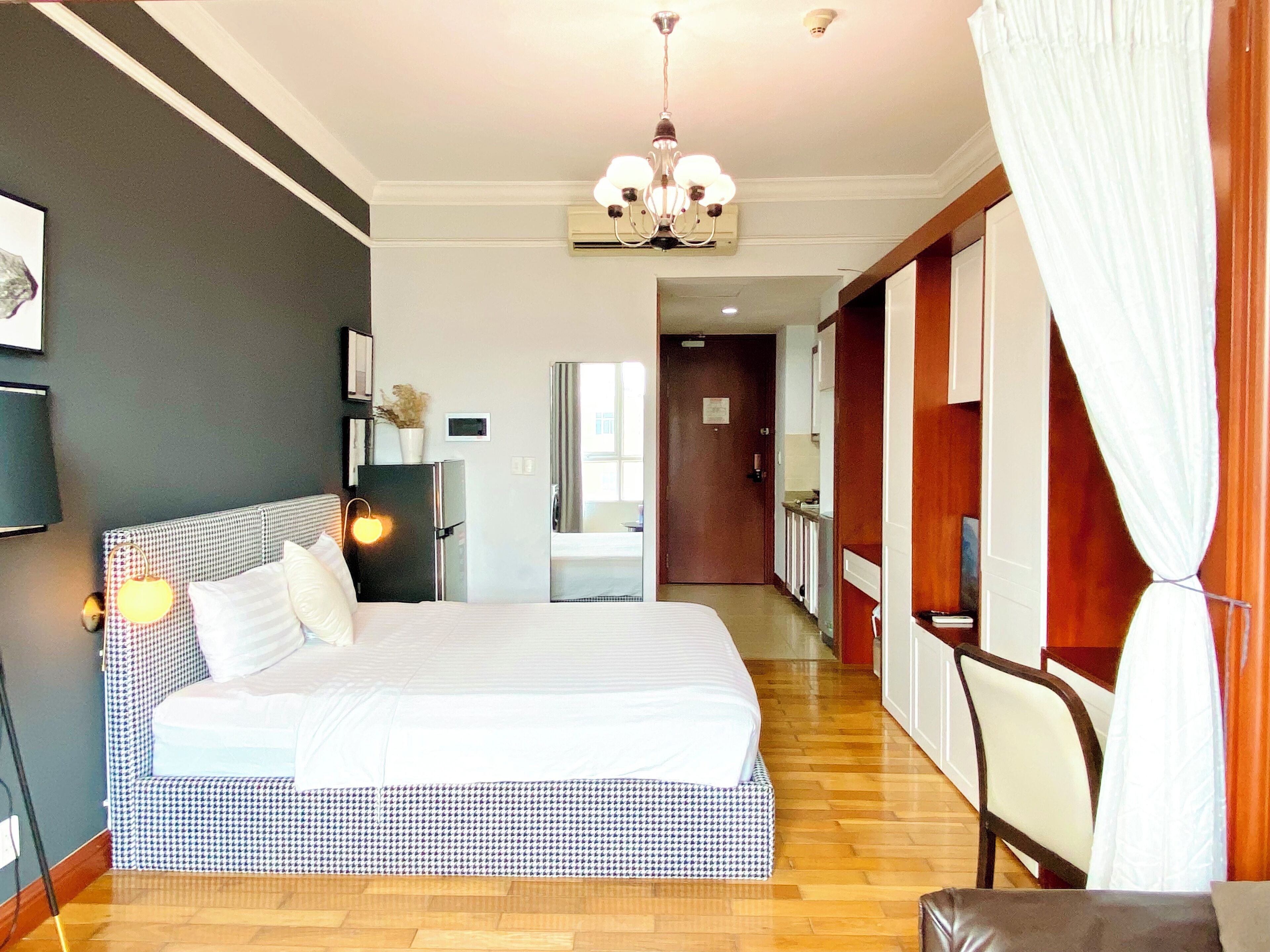 A hotel room with a large bed, a chandelier, a refrigerator, and a view of the hallway.