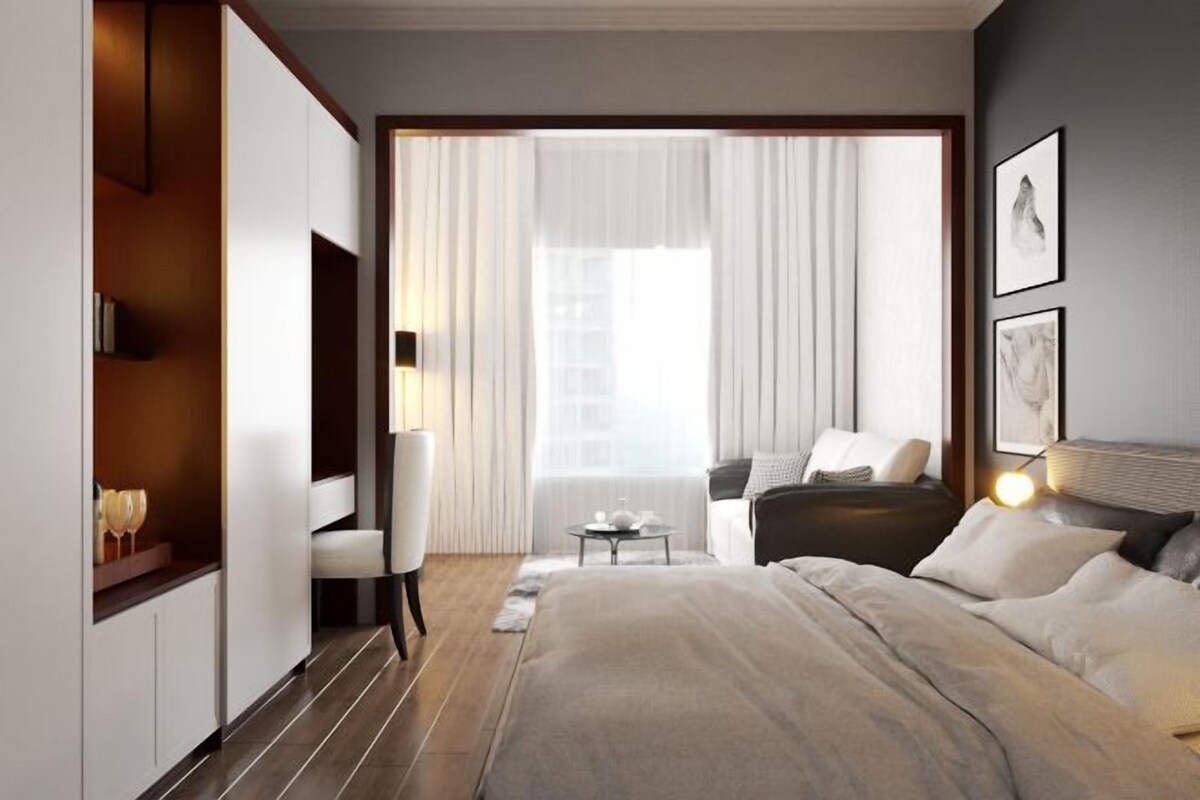 A modern hotel room with a large bed, a desk, and a chair. There is a window with curtains and a view of the city.