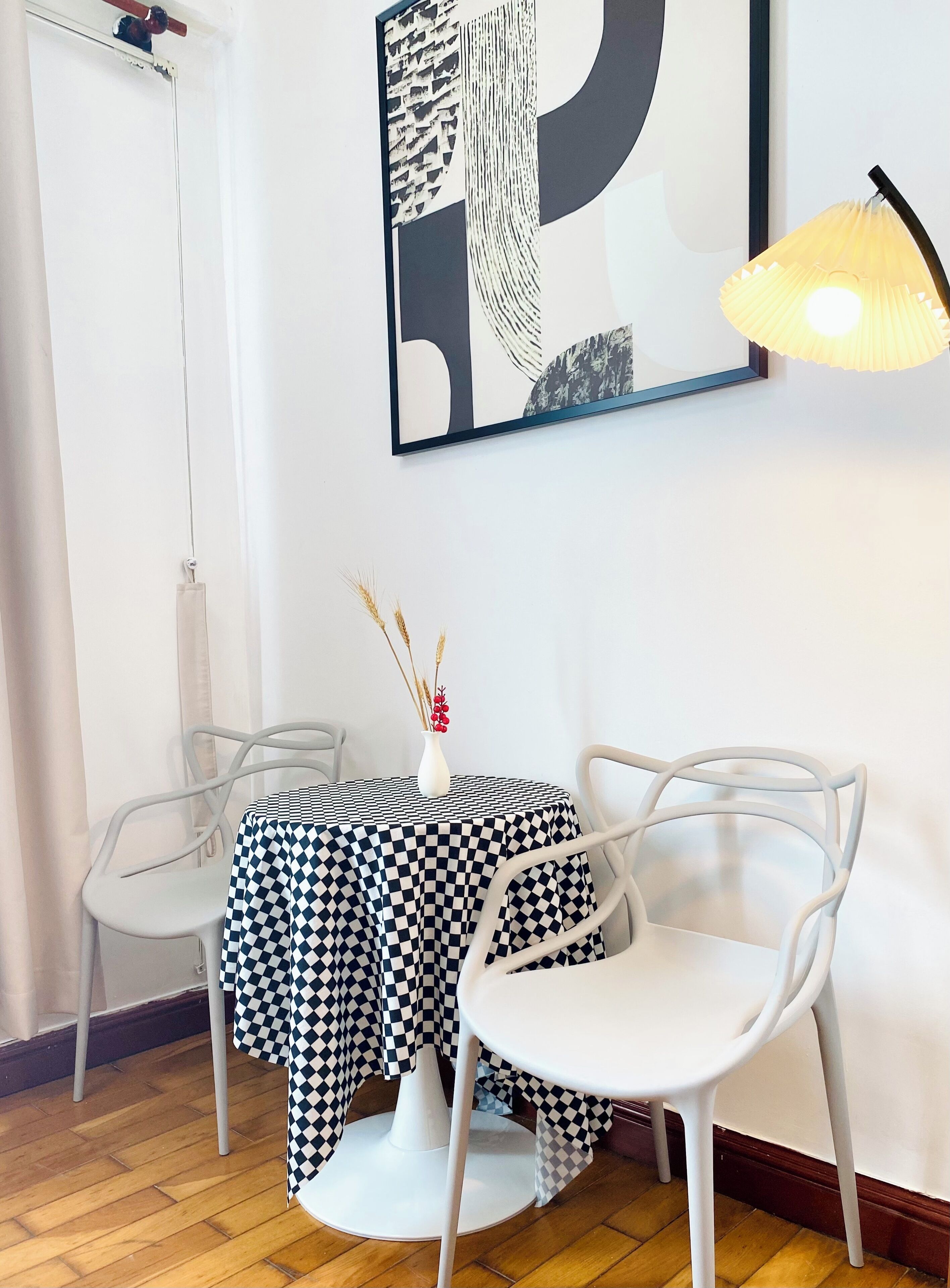 A dining area with a checkered tablecloth, two white chairs, a vase with dried plants, and a large abstract painting on the wall.