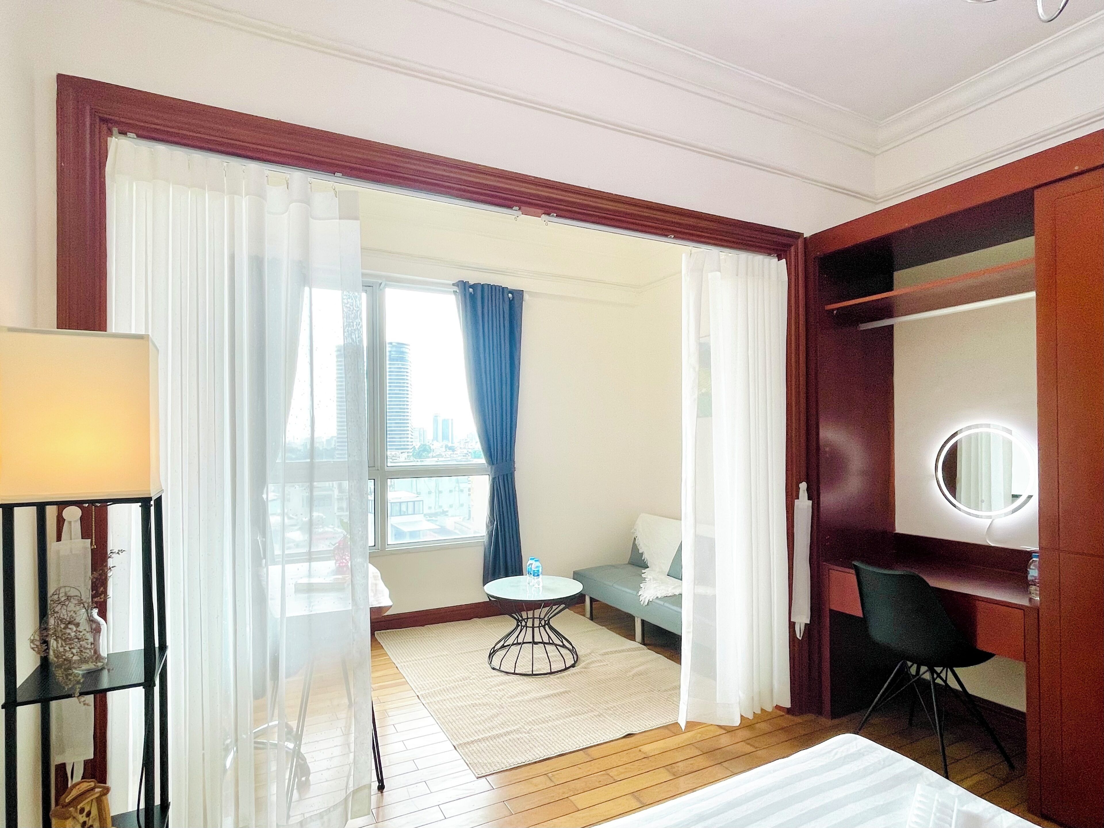 Una habitación de hotel con una cama, un espejo redondo, una mesita, una silla, un sofá y vistas de la ciudad a través de la ventana.