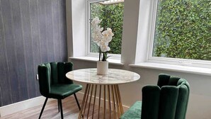 A room with a round table, two green chairs, a vase with white flowers, and a view of greenery through large windows. - Spacious 1-bed Studio Apt w/ Free Parking & WiFi (Bournemouth)