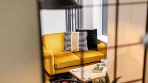 A yellow sofa with striped and black cushions, a wooden coffee table with a book and a plant, and a window with vertical blinds in the background. - NEW! Superb Studio Apt w/ Free Parking & WiFi (Bournemouth)