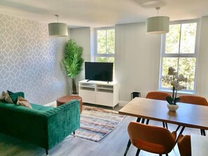 A modern living room with a green sofa, a dining table with orange chairs, a flat-screen TV on a white entertainment unit, and large windows with natural light.