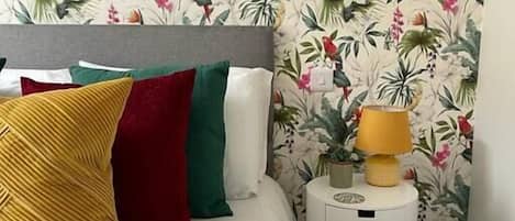 A bed with multiple pillows, a bedside table with a lamp, and a floral wallpaper pattern.
