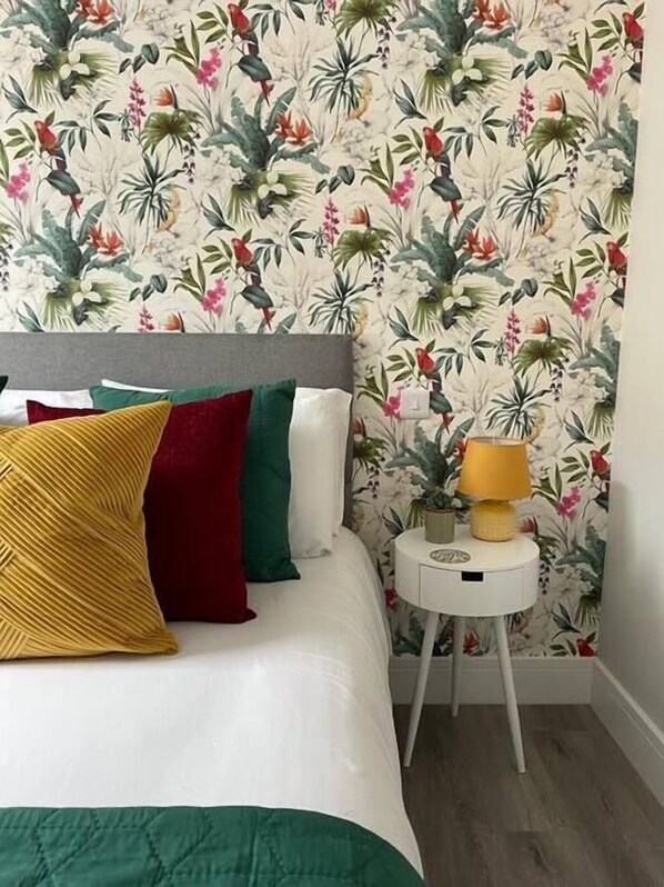 Una cama con varias almohadas, una mesita de noche con lámpara y un estampado floral en el papel tapiz.