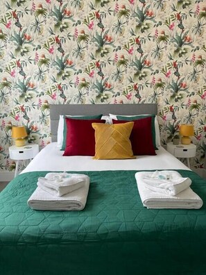 A neatly made bed with a green bedspread, two white towels, a red pillow, and a yellow pillow on a headboard.