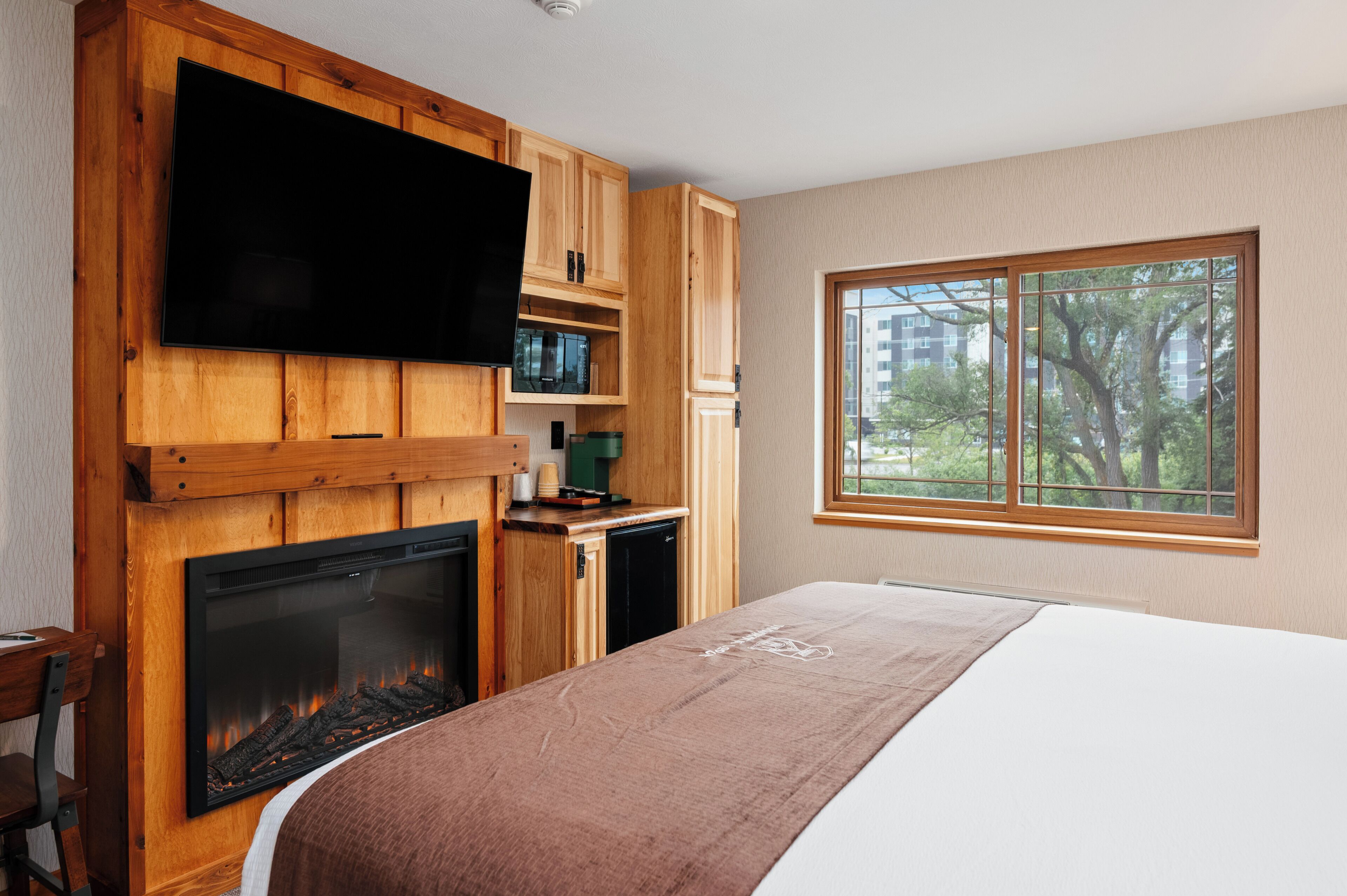 A bedroom with a fireplace, wooden cabinets, a television, and a window with a view of trees and buildings.