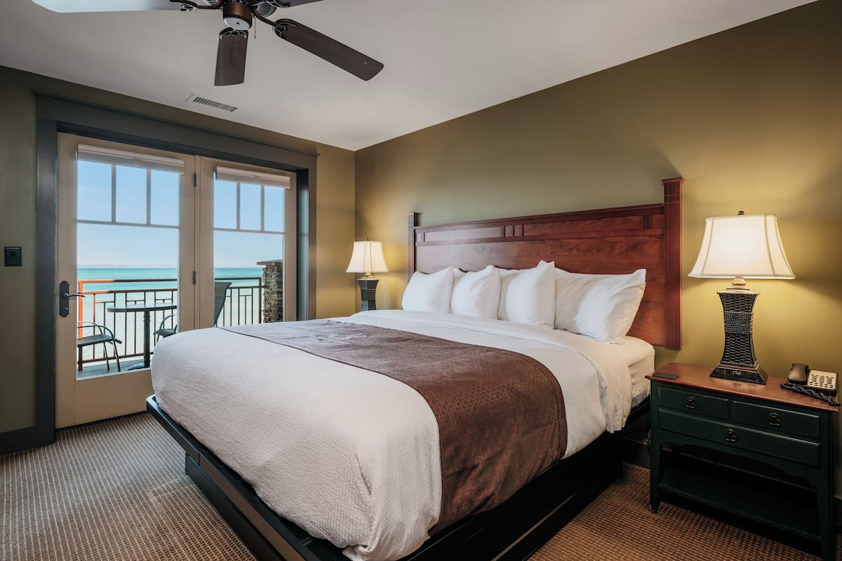 A hotel room with a bed, two bedside lamps, a nightstand, and a view of the ocean through a sliding door.